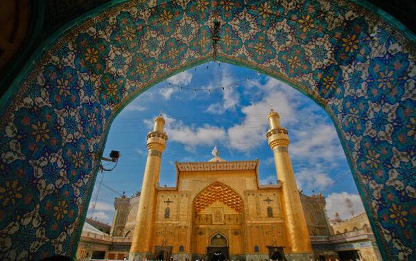 View through a blue and ornate archway of the golden Imam Ali shrine in Najaf, Iraq, with two minarets and a blue sky with clouds. Keywords: Imam Ali shrine, Najaf, Iraq, secret burial, history of construction, architectural features, imamate, pilgrimage, Muslim community. Donate crypto via ETH for Islamic relief.