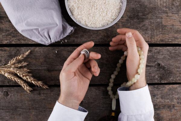 Hands holding coins and prayer beads near rice and wheat, symbolizing contemplation of wealth calculation and charity, with crypto donations like BTC accepted.