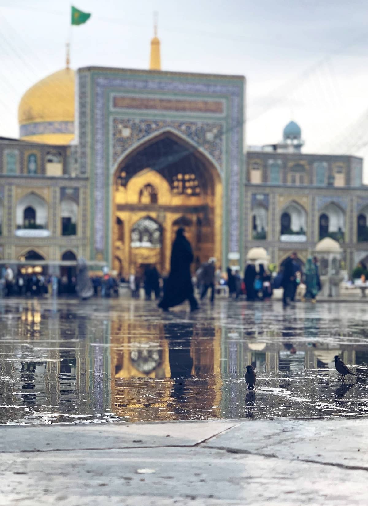Golden domed shrine with ornate entrance and people walking in the rain, reflections on wet ground. Crypto donation via XRP.