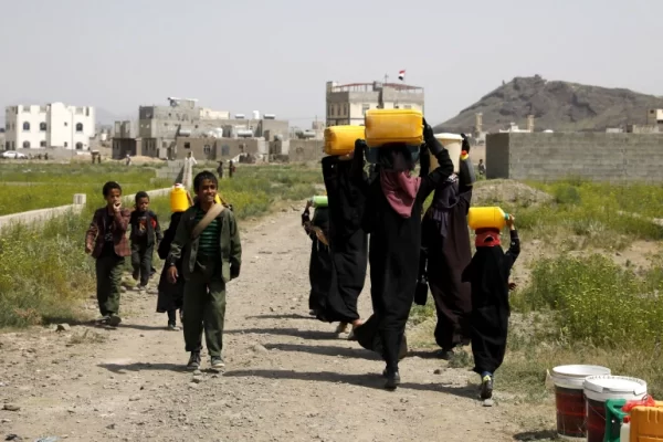 Children and women carrying water containers on their heads in a dusty village, with buildings in the background. Aid supported by Arab Emirates Dirham.