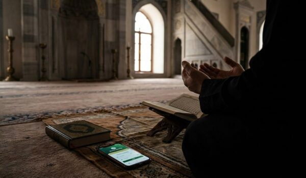 Person in prayer with hands raised in a mosque, with Quran and phone showing donation app, performing spiritual retreat and seeking Laylatul Qadr, donating via crypto and stablecoins like USDC.