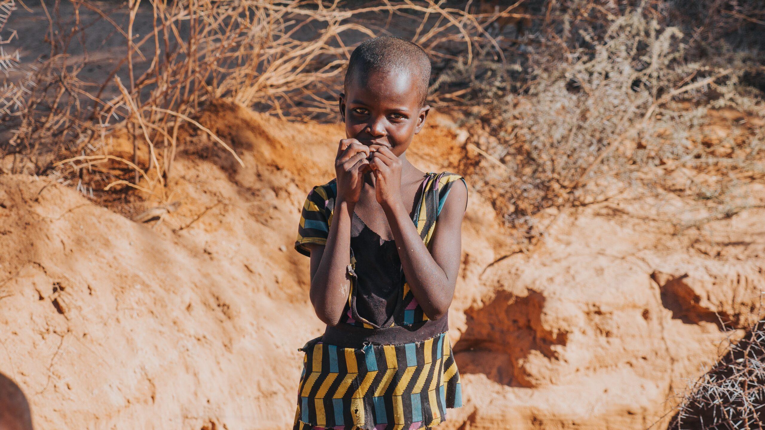 A young African child with hopeful eyes, her hands clasped near her face, stands against a dry, sandy backdrop. The image implies a need for aid, emphasizing crypto donation support via SOL.